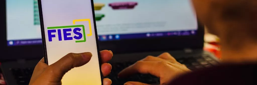 moça segurando celular com app do fies e usando computador